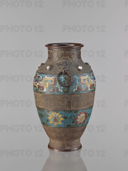 Vase, Chinese, Qing dynasty 1644–1911, 18th century, China; porcelain vessel decorated with lotuses, plant motifs including lotus tendril ornament, dragons, and taotie; height 30.5 cm., An ornate vase sits against a plain light gray background, showing a bulbous body with a narrow neck and flared rim. The vase surface combines metallic bronze tones with bands of bright turquoise blue and accents of gold, red, white, green, and brown. A decorative metal ring handle is attached near the shoulder, framed by raised ornamental motifs and small round insets. Patterned bands encircle the vessel, including a central turquoise band with stylized floral forms in gold and red and upper and lower bands of densely textured geometric surface in bronze. The rim and base show darker brown metallic finishes, and the overall surface has a slightly aged patina. The vase rests on a reflective surface that mirrors its lower portion.