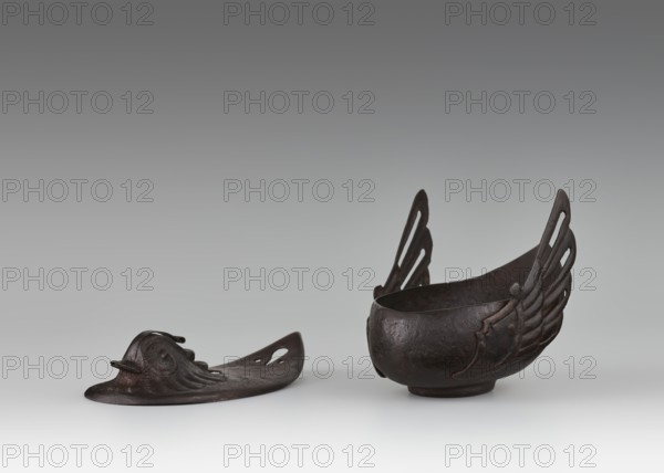 Duck incense burner, Chinese, Qing dynasty 1644–1911, 19th century. Bronze vessel in the form of a duck with wings, function: incense burner. Dimensions: height 9.7 cm; width 13.6 cm. Subjects: ducks, birds. Provenance unknown., A dark brown metal object resembling a shallow bowl with tall wing-like decorative elements on opposite sides sits next to a separate flat, elongated dark brown metal piece with raised sculptural detailing that suggests a stylized animal head and flowing forms; the objects rest on a smooth light gray surface against a gradient gray background, with visible colors including dark brown, light gray, and various shades of gray.
