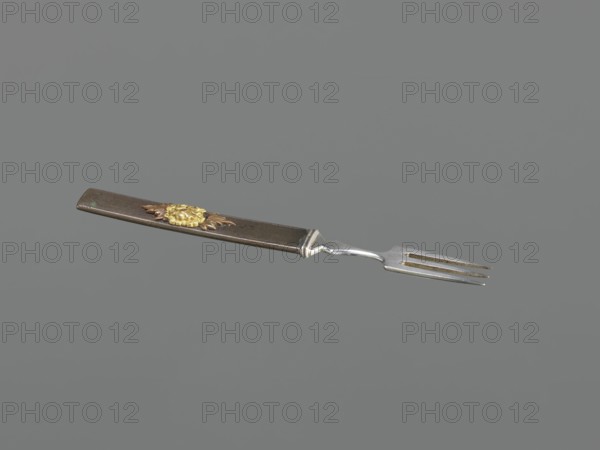 Fork with knife handle kozuka, Krüger, HB dr. Japanese, Edo period 1603–1868, 18th century. Melee weapons / sword accessory kozuka used with katana. Plastic decoration: chrysanthemum flower with two leaves; textured background. Materials: plastic decoration. Function: cutlery/kozuka. Dimensions: height 16.6 cm, width 1.4 cm, depth 0.8 cm., An image of a fork with a long rectangular handle and a three-pronged metal tine section, shown against a solid gray background. The handle appears dark brown with a textured surface and features a decorative floral ornament in gold and bronze tones near one end. The metal tines and connector are silver with some darker discoloration or patina. Overall colors visible include gray, brown, gold, bronze, and silver.