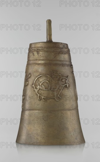 Bell, Persian, Qajar dynasty 1785–1925, 19th century, Iran; figurative motifs: animals—lions, camels, deer; height 26.5 cm, diameter 15.0 cm; Islamic art., A single metal bell-like object stands upright against a pale gray background. The object has a tapered cylindrical shape with a narrow top and a wider flared base, and a short vertical handle or knob protruding from the center of the top. The surface shows a textured, slightly pitted finish with visible wear and small scratches. A raised decorative motif wraps around the middle section, appearing as a stylized animal or spiral-like figure in low relief. The object displays a range of metallic tones including muted gold, bronze, and darker brownish areas where patina or shading is present. The lighting produces soft highlights and subtle reflections on the metal surface.