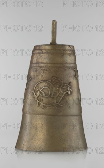 Bell, Islamic art, Persian, Qajar dynasty 1785–1925, figurative motifs: animals, lions, camels, deer, 19th century, Iran, varia; height 26.5 cm, diameter 15.0 cm, A single metallic bell-shaped object stands upright against a plain light gray background, showing a warm golden-bronze color with areas of darker patina and slight mottling. The surface displays textured wear and subtle scratches, with a raised decorative relief wrapping around the middle section depicting a stylized curled creature motif. A short vertical handle projects from the top, sharing the same bronze tones and aged finish. The lower flared portion of the bell is smoother and slightly shinier than the upper section, and a faint reflection appears beneath the object on the surface it rests on.