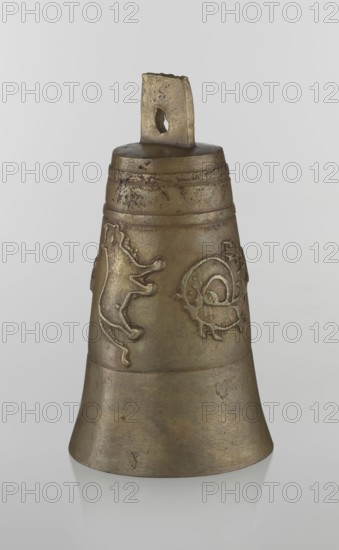 Bell, Islamic art, Persian culture, Qajar dynasty 1785–1925, figurative motifs: animals, lions, camels, deer, 19th century, Iran, varia; height 26.5 cm, diameter 15.0 cm, The image shows a single metal bell with a tapered cylindrical shape and a flat top with a rectangular loop handle that has an irregular hole through it. The bell's surface is textured and worn with raised decorative motifs on the upper section including a stylized animal-like figure and a circular ornamental design. The metal has a patinated appearance with shades of brown, bronze, and muted gold, along with darker areas of tarnish and subtle greenish undertones. The background is a smooth pale gray, and the bell casts a faint reflection on the glossy surface beneath it.