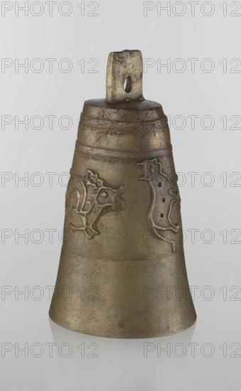 Bell, Islamic art, Persian, Qajar dynasty 1785–1925, figurative motifs: animals, lions, camels, deer; 19th century, Iran; height 26.5 cm, diameter 15.0 cm, The image shows a single metallic bell standing upright against a pale gray background, with a slightly reflective surface beneath it. The bell is a muted bronze color with areas of darker brown patina and subtle greenish tints, and it has a worn, textured surface. Near the top there is a rectangular loop with a hole, showing rough, uneven edges and darker discoloration. The bell's body tapers outward toward the base and features a few horizontal raised bands and a few raised decorative motifs in a similar bronze tone with slightly lighter highlights. The surface exhibits scratches, small pits, and areas of uneven coloration, and the base edge shows faint reflections on the smooth surface below.