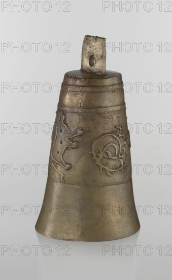 Bell, Persian, Qajar dynasty 1785–1925, 19th century, Iran; figurative motifs: animals lions, camels, deer; material/technique: unspecified; height 26.5 cm, diameter 15.0 cm., A single metallic bell with a worn surface sits upright against a pale gray background, showing a mottled bronze and brown coloration with areas of darker patina; the bell has a slightly flared base and a narrower top section with a rough rectangular loop for hanging, which has a hole through it and shows pitting and irregular edges; a few raised decorative motifs appear around the upper body, including a stylized animal-like shape and a swirling organic form, both matching the bell’s bronze tones but outlined by subtle shadows and highlights; the surface shows scratches, small dents, and uneven texture, and the bell casts a faint reflection on the glossy surface beneath it.