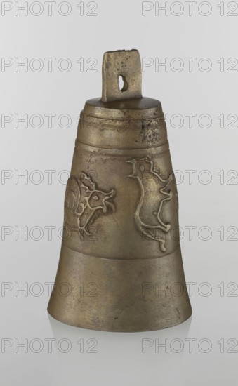 Bell, Islamic art, Persian, Qajar dynasty 1785–1925, figurative motifs, animals, lions, camels, deer, 19th century, Iran, varia; height 26.5 cm, diameter 15.0 cm, A single metal bell with a patinated surface in shades of brown, bronze, and muted gold sits against a plain light gray background; the bell has a rectangular loop at the top with a rounded hole and shows surface wear and darker speckles, a midsection with raised decorative animal-like reliefs in similar brown and gold tones, a banded upper portion with faint horizontal ridges, and a flared lower skirt with subtle scratches and uneven coloration.
