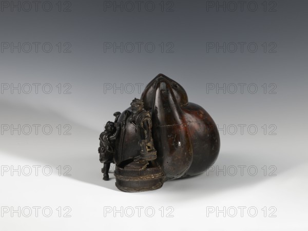 Censer kôro, figurative scenes, Japanese art, gourds, Japanese culture, Meiji 1868–1912, bronze. Decorated with figurines of Japanese people busy scrubbing its surface. Incense burner in the shape of a leather cistern, 2nd half of the 19th century, Japan; vessel, censer; height 16.3 cm, width 26.5 cm., A dark brown, glossy carved object with a rounded, bulbous form sits on a pale surface against a gradient gray background; a carved figure in similar dark brown material stands beside the rounded form, connected to a pedestal-like base and accompanied by a smaller carved element with curled details, and the object shows subtle variations of brown and black with some lighter brown patches and worn areas.