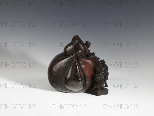 Censer kôro, figurative scenes, Japanese art, gourds, Japanese culture, Meiji 1868–1912, bronzes. Decorated with figurines of Japanese people busy scrubbing its surface. Incense burner in the shape of a leather cistern, 2nd half of the 19th century, Japan; vessel, censer; height 16.3 cm, width 26.5 cm., A dark brown glossy sculpted object with rounded, bulbous shapes sits slightly right of center on a light surface, casting a soft shadow to the left; the sculpture has smooth, curving forms with a few subtle indentations and a worn, slightly reddish-brown area on one side. On the right side of the main form, a small detailed figure in a darker brown hue stands on a square base, with intricately carved folds and raised elements. The background transitions from a pale off-white at the bottom to a muted gray at the top, and the overall color palette includes shades of dark brown, reddish-brown, gray, and off-white.