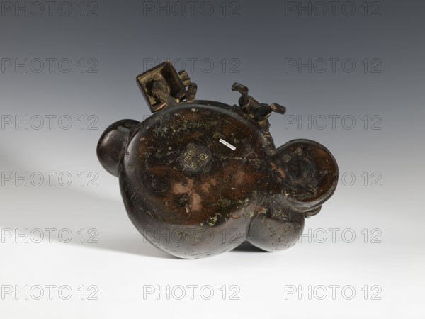 Censer koro, figurative scenes, Japanese art, gourds, Japanese culture, Meiji period 1868–1912, bronze. Decorated with figurines of Japanese people busy scrubbing its surface. Incense burner in the shape of a leather cistern, 2nd half of the 19th century, Japan; vessel censer; height 16.3 cm, width 26.5 cm., A dark brown, irregularly rounded object with a glossy, mottled surface sits on a light surface against a gradient gray background; the object has a few bulbous protrusions and a small square section attached near the top with sculpted, angular elements extending from it, and the surface shows patches of lighter brown and greenish discoloration and textured wear.