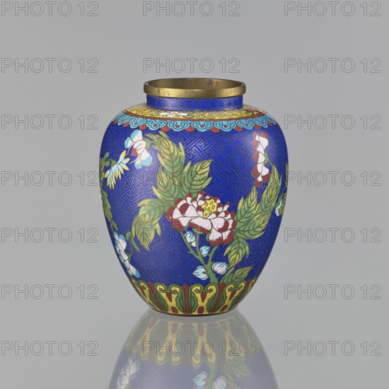 Vase, cloisonné enamel, Chinese, oval-shaped with a short neck and ring-shaped spout, without a foot; decorated on the central belly with colorful peonies on a sapphire background and a Chinese meander motif; 19th century Qing, 1644–1911; height 16.5 cm, diameter 13.8 cm; maker, provenance unknown., A rounded vase with a narrow neck and a metallic rim sits against a plain gray background, its surface predominantly bright blue with an intricate darker blue geometric pattern. The body is decorated with floral motifs in green leaves and blossoms showing white, pink, red, and yellow tones, with small areas of light blue and pale turquoise in some flower accents. A band near the top features a scalloped cloud-like design in light blue and gold, and the base area shows a decorative border in green, yellow, and red. The vase casts a soft reflection on the reflective surface beneath it, mirroring the blue, green, yellow, red, and metallic colors.