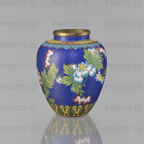 Vase, cloisonné enamel, Chinese, oval-shaped with short neck and ring-shaped spout, without a foot; decorated on the central belly with colorful peony flowers on a sapphire background and a Chinese meander motif; Qing dynasty 1644–1911, 19th century; height 16.5 cm, diameter 13.8 cm;, provenance unknown., A rounded decorative vase with a bright blue background featuring floral designs in green, white, red, yellow, and pale blue, a narrow neck with a metallic rim in a brass color, a patterned band near the top in yellow and turquoise, leafy green foliage surrounding several white and pale blue flowers with yellow centers, additional red and white blossoms, and a decorative green and yellow border near the base; the vase sits on a reflective gray surface that mirrors its colors.