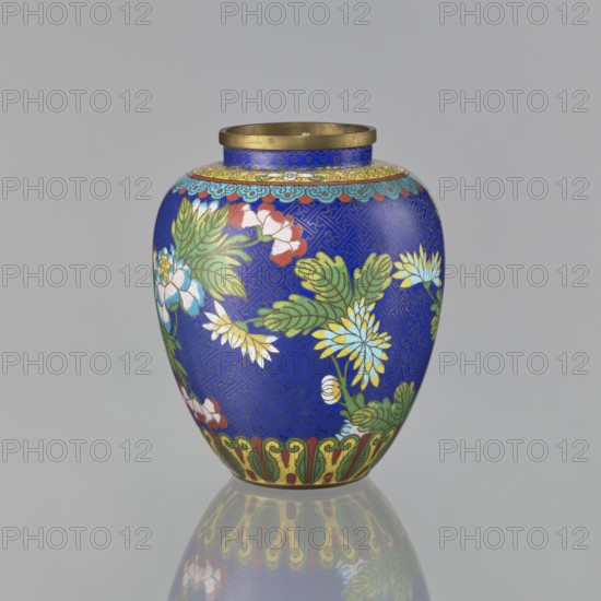 Vase, cloisonné enamel, Chinese art; plant ornament featuring peonies and a meander motif; provenance, Qing dynasty 1644–1911, 19th century; oval-shaped vase with a short neck and ring-shaped spout, without a foot; decorated on the central belly with colorful flowers on a sapphire background and additional Chinese meander motif; China; vessel; height 16.5 cm, diameter 13.8 cm., A rounded decorative vase sits against a plain light gray background, its main surface a vivid deep blue patterned with an intricate geometric design. The vase is adorned with painted floral motifs in green, yellow, white, pale blue, red, and pink, with leaves in various shades of green and flower centers in yellow. A band near the top features a scalloped cloud-like border in light blue and teal over a yellow background, while the rim is metallic gold. The base area shows a stylized ornamental border in green, yellow, and red, and the vase casts a soft mirrored reflection on the glossy surface beneath it.