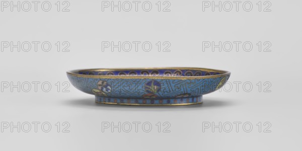 Bowl, gift; Chinese, Qing 1644–1911; cloisonné enamel; 18th century, China; vessel, diameter 9.3 cm. Shallow bowl on a separate foot decorated inside with a colorful composition of three symbolic fruits: a peach, a pomegranate and a finger lemon, accompanied by bats—interpreted as a wish for “many happiness, long life, and many sons.” Inner rim decorated with a circle of ruyi heads black, red, navy blue. Outer surface filled with colorful medallions arranged singly, in pairs, or in threes, featuring stylized plant motifs framed by a Chinese meander pattern made of bent wire in a net-like form on a turquoise background. Foot decorated with a band of alternating red and blue circles on turquoise. Bottom entirely covered with turquoise enamel., A shallow decorative bowl sits on a flat surface, shown in profile with a low rounded rim and a narrow foot. The exterior and interior surfaces display intricate enamel patterns in blue tones, with accents of gold around the rim and foot. There are...