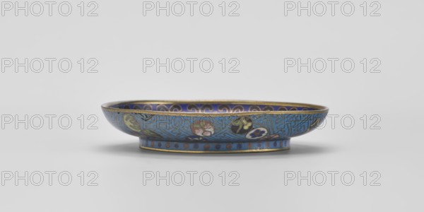 Bowl, cloisonné enamel, 18th century, China. Shallow bowl on a separate foot decorated inside with a colorful composition of three symbolic fruits — a peach, a pomegranate, and a finger lemon — accompanied by bats. Interpreted as a wish for many happiness, long life, and many sons. Inner rim decorated with a circle of ruyi heads black, red, and navy blue. Outer surface filled with colorful medallions arranged singly, in pairs, or in threes, featuring stylized plant motifs within a Chinese meander frame made of bent wire in a net-like pattern on a turquoise background. Foot decorated with a band of alternating red and blue circles on a turquoise ground. Bottom entirely covered with turquoise enamel. Diameter 9.3 cm., A shallow, round dish sits against a pale gray background, decorated in primarily blue tones with accents of gold, green, pink, black, white, and purple; the exterior shows a patterned blue field with circular medallions containing floral motifs in green, pink, black, and w...
