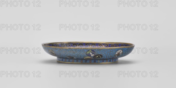 Bowl, cloisonné enamel, China, Qing 1644–1911, 18th century, diameter 9.3 cm. Shallow bowl on a separate foot decorated inside with a colorful composition of three symbolic fruits—peach, pomegranate, and finger lemon—accompanied by bats, interpreted as a wish for many happiness, long life, and many sons. Inner rim decorated with a circle of ruyi heads black, red, and navy blue. Outer surface filled with colorful medallions single, paired, or in threes featuring stylized plant motifs framed by a Chinese meander pattern made of bent wire in the form of a net, on a turquoise background. Foot decorated with a band of alternating red and blue circles on turquoise; bottom entirely covered with turquoise enamel.,  noted., A shallow decorative dish sits on a plain light surface against a pale background, showing a band of blue on its exterior with intricate line patterns in a lighter blue and metallic outlines, accented by several floral or petal-like motifs in colors including white, green, y...