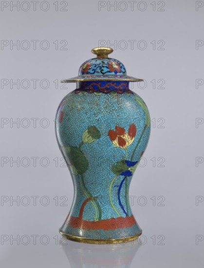 Vase with cover, cloisonné enamel baluster vase decorated with lotuses, Chinese, Qing dynasty 1644–1911, 18th century, height 37 cm., A lidded vase with a rounded body and flared base standing on a reflective surface, decorated overall in turquoise with an intricate maze-like pattern, featuring floral motifs including lotus flowers and leaves in red, yellow, green and blue. The lid has a domed shape with a small gold knob and a wide rim decorated in blue with red and gold accents. The vase shows gold trim along the base and rim, with additional red decorative banding near the bottom and green stems and leaves rising along the body. The background is a neutral pale gray.