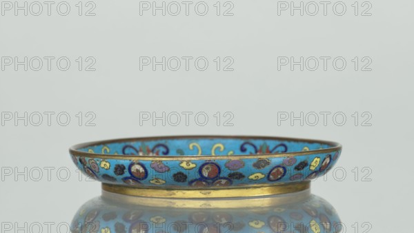 Plate, gift, Chinese, Qing dynasty 1644–1911, cloisonné enamel. Round plate with a flat bottom, without a distinct facet, with an arched rim on a low foot arranged in a ring at the rim. The mirror is decorated with a tondo surrounded by a multicolored lotus tendril. The composition in the tondo depicts a cricket in motion, perching on a flowering plant jumping from one flower to another, positioned on the right; flowering plants dominate the image. The outer rim is filled with medallions depicting stylized Chinese symbols lotus, peach, surrounded by cloud-like projections. The underside of the bottom is covered with single-color turquoise enamel. 19th century, China. Height 2 cm, diameter 14.7 cm., A shallow decorative dish with a bronze-colored rim and base sits on a reflective surface, the body of the dish showing a bright blue background decorated with a repeating motif of small cloud-like shapes and circular medallions in colors including blue, yellow, purple, black, red, and gold,...