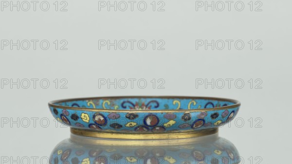 Round cloisonné enamel plate, 19th century, China. Round plate with a flat bottom, without a distinct facet, with an arched rim, on a low foot arranged in a ring at the rim. The mirror is decorated with a tondo surrounded by a multicolored lotus tendril. The composition enclosed in the tondo depicts an insect cricket in motion, perching on a flowering plant jumping from one flower to another; the insect is located on the right side of the composition. Flowering plants dominate the entire image. The outer rim is filled with medallions depicting stylized Chinese symbols lotus, peach, surrounded by free-form projections of clouds. The underside of the bottom is covered with single-color turquoise enamel. Height 2 cm; diameter 14.7 cm., A shallow round dish with a raised rim sits centered against a pale gray background, the dish painted primarily in bright turquoise blue with a band of decorative motifs around the rim in colors including red, yellow, purple, black, and dark blue, and edged...