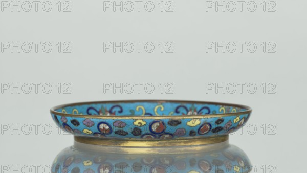 Plate, Chinese, Qing dynasty 1644–1911, 19th century. Cloisonné enamel round plate with a flat bottom, arched rim, on a low foot. Mirror decorated with a tondo surrounded by a multicolored lotus tendril; tondo depicts a cricket in motion perching on flowering plants, positioned at right, with flowering plants dominating the composition. Outer rim filled with medallions showing stylized Chinese symbols lotus, peach surrounded by cloud-like projections. Underside of the bottom covered with single-color turquoise enamel. Vessel; height 2 cm, diameter 14.7 cm., A shallow decorative bowl with a rounded rim sits on a reflective surface, showing a reflection beneath. The bowl has a bright blue background with patterns in colors including gold, yellow, red, purple, black, and dark blue, and a metallic gold band along its base and rim. The design includes small circular motifs and stylized cloud-like shapes in the various colors, and the interior shows similar blue and gold decorative elements....