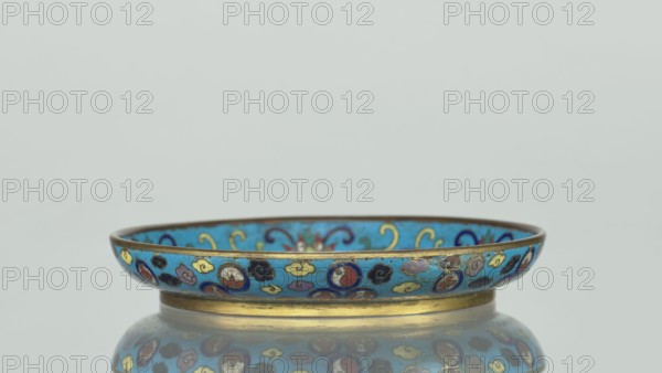Plate, cloisonné enamel, Round plate with a flat bottom, without a distinct facet, with an arched rim, on a low foot arranged in a ring at the rim. The mirror is decorated with a tondo surrounded by a multicolored lotus tendril. The composition enclosed in the tondo depicts an insect cricket in motion, perching on a flowering plant jumping from one flower to another. The insect is located on the right side of the composition. Flowering plants dominate the entire image. The outer rim is filled with medallions depicting stylized Chinese symbols lotus, peach, surrounded by free-form projections of clouds. The underside of the bottom is covered with single-color turquoise enamel. 19th century, China, vessel; height 2 cm, diameter 14.7 cm., A shallow bowl with a rounded rim sits on a reflective surface, showing a mirrored image beneath. The bowl has a bright turquoise-blue background with ornate painted motifs in colors including gold, dark blue, red, purple, black, green, and cream. The ri...