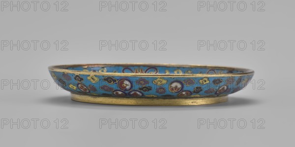 Plate, Chinese, Qing dynasty 1644–1911, 19th century. Cloisonné enamel round plate with a flat bottom, arched rim, on a low ring foot; height 2 cm, diameter 14.7 cm. Mirror decorated with a tondo surrounded by a multicolored lotus tendril. Tondo shows a cricket in motion perched on a flowering plant, positioned at left, with flowering plants dominating the scene. Outer rim filled with medallions depicting stylized lotus and peaches, surrounded by cloud-like projections. Underside of the bottom covered with single-color turquoise enamel., A shallow round dish with a low cylindrical foot, decorated all around with a turquoise blue background and a repeating pattern of small cloudlike motifs and circular medallions in colors including gold, dark blue, red, white, yellow, and purple, the rim and foot showing a metallic gold tone, set against a neutral light gray surface and background.