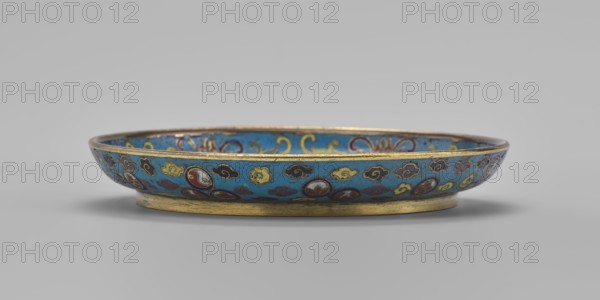 Round plate with a flat bottom, without a distinct facet, with an arched rim, on a low foot arranged in a ring at the rim. Cloisonné enamel, 19th century, Qing dynasty, China. The mirror is decorated with a tondo surrounded by a multicolored lotus tendril. The composition in the tondo depicts an insect cricket in motion, perching on a flowering plant jumping from one flower to another, positioned on the left; flowering plants dominate the image. The outer rim is filled with medallions depicting stylized Chinese symbols lotus, peach, surrounded by free-form cloud projections. The underside of the bottom is covered with single-color turquoise enamel. Vessel; height 2 cm, diameter 14.7 cm., inscription/markings not specified., A shallow decorative dish shown in profile with a low foot and a gently flared rim, the exterior and interior surfaces covered in a bright blue enamel field decorated with a repeating pattern of stylized cloud- and rosette-like motifs in yellow, dark blue, red, and ...