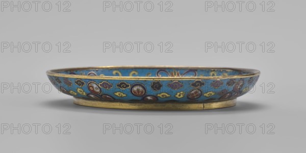 Plate, Chinese, Qing dynasty 1644–1911, cloisonné enamel, 19th century. Round plate with a flat bottom, without a distinct facet, with an arched rim on a low foot arranged in a ring at the rim; height 2 cm, diameter 14.7 cm. The mirror is decorated with a tondo surrounded by a multicolored lotus tendril. The composition in the tondo depicts a cricket in motion perching on a flowering plant jumping from one flower to another, the insect located on the left side; flowering plants dominate the image. The outer rim is filled with medallions depicting stylized Chinese symbols lotus, peach, surrounded by free-form cloud projections. The underside of the bottom is covered with single-color turquoise enamel., A shallow round dish sits on a low circular foot, showing a bright turquoise interior and exterior background decorated with a repeating pattern of round medallions and cloudlike motifs in colors including deep blue, dark red, yellow, purple, white, and black, with metallic gold edging ar...