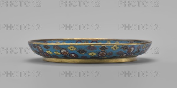Round cloisonné enamel plate, Qing dynasty, 19th century, China. Round plate with a flat bottom, without a distinct facet, arched rim on a low ring foot; height 2 cm, diameter 14.7 cm. Mirror decorated with a tondo surrounded by a multicolored lotus tendril; the tondo shows a cricket in motion perching on a flowering plant, positioned at the left, with flowering plants dominating the composition. Outer rim filled with medallions depicting stylized lotus and peach symbols, surrounded by cloud-like projections. Underside of the bottom covered with single-color turquoise enamel., A shallow round dish with a low foot, shown in profile, resting on a flat surface; the dish has a bright turquoise-blue background decorated with a repeating pattern of cloud-like motifs and round medallions in colors including yellow, navy, maroon, white, and black, with thin metallic gold outlining around the rim and along the base; the surface shows subtle variations in color and a soft shadow beneath the dish...