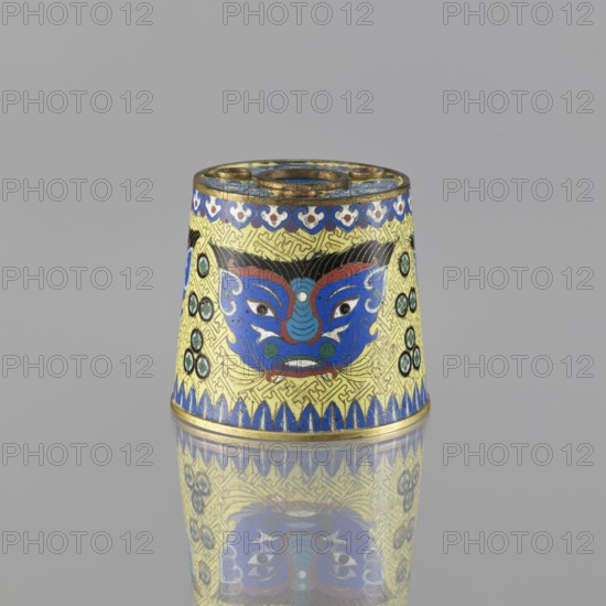 Sawdust container, gift; Chinese, Qing dynasty 1644–1911; cloisonné enamel vessel with beasts fantasy, tiger symbol, meander ornament; base with four inserts; decoration: on a yellow background, four masks against a grid background; 19th century, China; height 8.5 cm, diameter 9.7 cm., A small conical metal object with a decorative mask motif dominates the center, featuring bold blue, red, black, green, gold, and white enamel. The body shows a repeated geometric background in gold with a blue and gold scalloped border along the top and a blue pointed border along the bottom. On the central face-like design, blue is prominent with red accents and black detailing, and green circular insets flank the sides. The top rim is metallic gold with small recessed sections, and the entire object sits on a reflective grey surface that mirrors its colors and patterns.