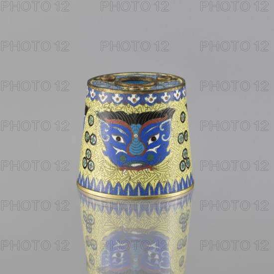 Sawdust container, Cloisonné enamel vessel, Qing dynasty 1644–1911, 19th century, China. Base with four inserts. Decoration on a yellow background: four masks against a grid background; motifs include beasts fantasy, tiger symbol, and meander ornament. Height 8.5 cm; diameter 9.7 cm., A small conical object sits against a plain gray background, its surface decorated with a bright yellow base and intricate patterns in blue, black, red, green, and gold; a stylized blue face with white eyes and red accents is centered on the visible side, flanked by circular green and black medallions, and a band of blue petal-like shapes runs along the lower edge, while a decorative blue and white border encircles the top; the object casts a clear reflection on the glossy surface beneath it.