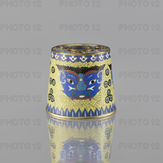 Sawdust container, Paderewski, Ignacy Jan 1860-1941, Chinese art, Chinese culture, Qing 1644–1911, cloisonné enamel, beasts fantasy, tiger symbol, meander ornament. Base with four inserts. Decoration: on a yellow background, four masks against a grid background. 19th century, China. Vessel; height 8.5 cm, diameter 9.7 cm., A conical decorative object sits on a reflective surface, showing a reflection below. The object features a dominant yellow background with an intricate repeating pattern and prominent blue, red, white, green, black, and gold decorative elements. A stylized face motif in blue with red and white accents appears on the visible side, flanked by circular green and black roundels on both sides. The top rim and base are accented in gold with blue decorative borders, and the top surface has circular openings and gold detailing. The overall scene includes the gray backdrop behind the object.