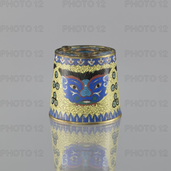Sawdust container, Chinese, Qing dynasty 1644–1911, cloisonné enamel, decorated with fantasy beasts, meander ornament, tiger symbol, base with four inserts; on a yellow background, four masks against a grid background. 19th century, China; vessel; height 8.5 cm, diameter 9.7 cm, A small conical metal object sits upright on a reflective surface, showing a mirrored image below. The object is decorated with a central stylized face motif in bright blue with red and teal accents and black hair details, set against a textured yellow background with interlocking geometric patterns. A band of blue and gold decorative shapes runs along the top edge and a row of blue triangular shapes borders the bottom edge. Several circular enamel insets in shades of teal and black appear on the sides. The top rim and base have visible metallic gold tones, and the overall color palette includes blue, yellow, gold, black, red, and teal.