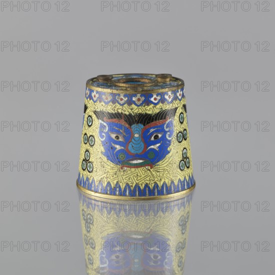 Sawdust container, gift; Chinese, Qing dynasty 1644–1911; cloisonné enamel; 19th century. Vessel with base with four inserts; height 8.5 cm, diameter 9.7 cm. Decoration: on a yellow background, four masks against a grid background; motifs include beasts fantasy, meander ornament, and tiger symbol., A small conical metal object with a brightly colored decorative surface sits on a reflective grey surface, showing a mirrored reflection below. The object is primarily yellow with extensive blue detailing and accents of red, green, black, and gold. A stylized face or mask motif in blue and red appears on the front, with black hairlike lines and green circular accents to its sides. The top rim and base edges have blue and gold bands with patterned designs, and there are repeated circular green-and-black elements on the sides. The overall surface shows intricate patterned background lines in gold and fine detailing in blue, red, green, black, and gold.