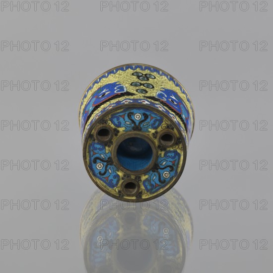 Sawdust container, gift, Chinese, Qing dynasty 1644–1911, cloisonné enamel vessel decorated with fantasy beasts, meander ornament, and tiger symbol; base with four inserts. Decoration: on a yellow background, four masks against a grid pattern. 19th century, China. Height 8.5 cm; diameter 9.7 cm., A cylindrical decorative object lies horizontally against a neutral gray background, showing its circular end facing the viewer and a reflective surface beneath that creates a mirrored image. The object has intricate enamel-like patterns in vivid blue, yellow, turquoise, black, white, red, and green, with stylized floral and abstract motifs wrapping around its body. The visible end has a central hollow opening surrounded by a ring and several smaller circular holes, all framed by ornate scrolling designs in blue and black on a yellow background with small white accents. The upper band shows repeating decorative elements in blue and yellow with touches of red and green, and the metal edges have...