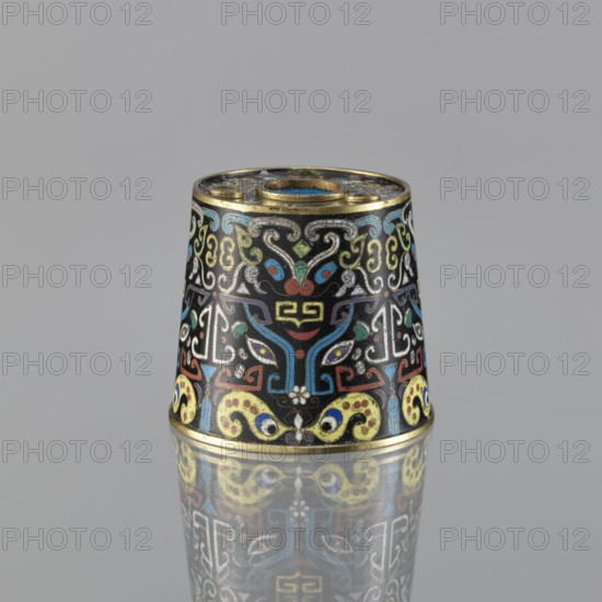 Sawdust container, Chinese, Qing dynasty 1644–1911, 19th century, cloisonné enamel vessel with beasts fantastical creatures and cicada motifs, base with four inserts, height 8.5 cm, diameter 9.7 cm., A small conical object sits centered on a reflective surface, showing a mirrored image beneath; the object has a black background richly decorated with ornate symmetrical patterns in colors including gold, blue, red, yellow, green, white, and turquoise, with gold rims at the top and bottom; the surface shows scrolling shapes, stylized motifs, and curved lines forming a repeating decorative design against a plain gray backdrop.