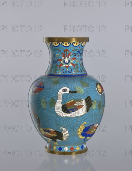 Vase, Cloisonné enamel vessel on a foot with a neck. Decoration: ducks on a turquoise background; lotus tendril on the neck. Chinese; Qing dynasty 1644–1911; 19th century. Function: decorative vessel. Materials: cloisonné enamel. Subject: ducks. Height: 23.5 cm., A rounded vase with a narrow neck sits against a plain light gray background; the vase is predominantly bright turquoise blue with swirling concentric line patterns and features a decorative band near the base in alternating diamond shapes of red, blue, yellow, white, and green, edged with metallic gold trim. The body of the vase is decorated with a group of stylized waterfowl rendered in white, black, brown, blue, red, and gold, and surrounded by green lotus leaves and curling gold and blue lines. The flaring neck displays an ornate floral motif in red, white, blue, yellow, and gold on the same turquoise background, with a gold rim at the top and a gold ring at the base reflected faintly on the surface below.