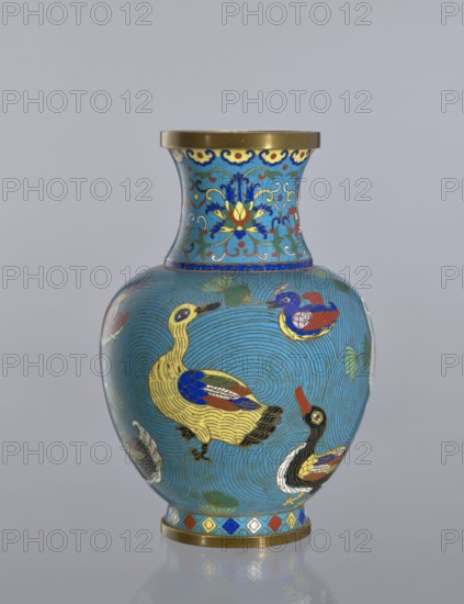 Vase, Cloisonné enamel vase on a foot with a neck. Decoration: ducks on a turquoise background; lotus tendril on the neck. Chinese, Qing dynasty 1644–1911, 19th century. Materials: cloisonné enamel. Subject: ducks. Height: 23.5 cm., A ceramic vase with a rounded body and flared neck sits against a pale gray background; the vase is predominantly bright turquoise blue with swirling line patterns and is decorated with stylized birds in shades of yellow, gold, blue, red, black, and white, along with small leaf motifs in green. The neck features a floral-like ornamental pattern in blue, yellow, red, and gold, with a gold rim at the top. A band of geometric shapes in blue, red, yellow, and white and a gold base ring appear at the bottom, and the vase casts a subtle reflection on the surface beneath it.