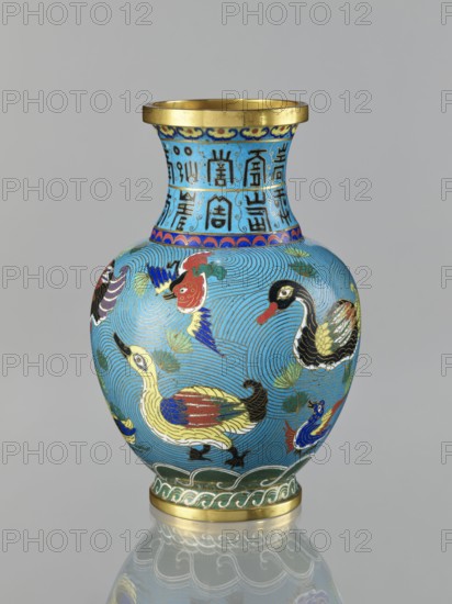 Vase, Chinese, Qing 1644–1911. Cloisonné enamel vase on a foot with a neck. Decoration: ducks on a turquoise background and an archaizing longevity shou inscription on the neck. 19th century, China. Height 23.3 cm., A rounded vase with a flared neck sits against a plain gray background, predominantly light blue with a gold rim at the top and a gold band at the base; the neck features a decorative band of darker blue squares containing vertical dark markings, and the shoulder below has a narrow band with alternating blue and red scallop shapes. The main body is covered in swirling line patterns in darker blue and decorated with a group of stylized birds in colors including yellow, red, black, white, green, and blue, with feather details in gold and dark outlines; there are also green leaf-like motifs and a green wavy band above the gold base. The vase sits on a reflective surface that creates a faint mirror image of the bottom portion.