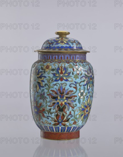 Vase with lid, Chinese, Qing dynasty 1644–1911, cloisonné enamel decorative vessel with floral ornaments and lotus tendril motifs, 18th–19th century, China; height 23 cm, diameter 14 cm., A lidded decorative jar sits against a plain light gray background, featuring a rounded body and a domed lid topped with a small knob. The jar displays an intricate floral pattern in a palette of blue, turquoise, red, green, white, black, gold, and purple. The main ground color appears turquoise with a mottled texture, while detailed flower motifs and scrolling vines cover the surface in shades of blue, red, green, white, black, and gold. The rim and lid edge show metallic gold accents, and the base band is a reddish color with gold ornament. The domed lid mirrors the jar’s decorative scheme with blue and gold patterns and a gold knob. The jar rests on a reflective surface that produces a soft mirror image below it.