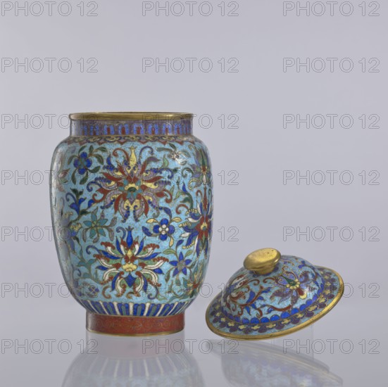 Vase with lid, Chinese, Qing dynasty 1644–1911, cloisonné enamel vessel with floral and plant ornaments including floral tendrils and lotus tendril motifs, decorative flowers; 18th–19th century, China; height 23 cm, diameter 14 cm., An ornate lidded jar and its lid sit against a pale gray background; the jar has a rounded body with a narrow rim and a separate domed lid positioned on its side nearby. Both pieces display an intricate floral pattern on a light blue ground, featuring flowers and scrolling foliage in colors including dark blue, red, green, and gold, with gold outlining and accents throughout. The rim and foot of the jar and the knob and edge of the lid show metallic gold tones, and there is a band of deep blue decoration near the base. The surface has a glossy finish that produces subtle reflections on the smooth tabletop, and the overall color palette includes light blue, dark blue, red, green, and gold.