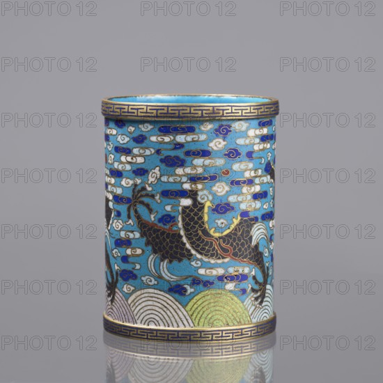 Brush vessel, cloisonné enamel, Qing dynasty 1644–1911, 19th century China. Decorated with dragons, clouds, waves/sea waves, claws, and meander ornament; turquoise interior. Height 12.7 cm; diameter 9.8 cm., A cylindrical decorative object with a predominantly turquoise blue background, decorated with a pattern of cloud-like shapes in white, dark blue, and gold. A large stylized creature with dark brown or black scaled body and gold outlines appears across the surface, with flowing tail and fins accented in white and gold. The top and bottom rims feature a geometric band in dark blue and gold. Near the base there are rounded wave-like motifs in pale green, white, and gold. The overall color palette includes turquoise blue, dark blue, white, gold, dark brown or black, and pale green.