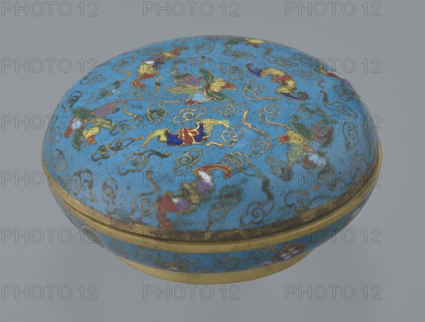 Round box with lid, cloisonné enamel, Chinese, Qing dynasty 1644–1911, 19th century. Cylindrical flattened shape on a separate ring foot with a slightly convex lid. Entire surface decorated with multi-colored lotus tendrils on a turquoise background. Lid shows three freely placed fruits — a peach, a pomegranate, and a lemon — accompanied by bats; motif interpreted as wishing many happiness, long life, and many sons. Asymmetrical composition set against cloud forms made of bent wire; same motif appears in the box's interior storage section, which is also decorated with colorful medallions single, paired, or grouped in threes featuring various Chinese plant ornaments. Rounded edges of lid and body, lower edge of foot and bottom are gilded. Bottom and entire interior surface covered with turquoise enamel. Container. Height 4.6 cm; diameter 9.8 cm., A rounded lidded container with a domed top and a circular base, predominantly bright blue in color with a gold rim and gold decorative outlin...