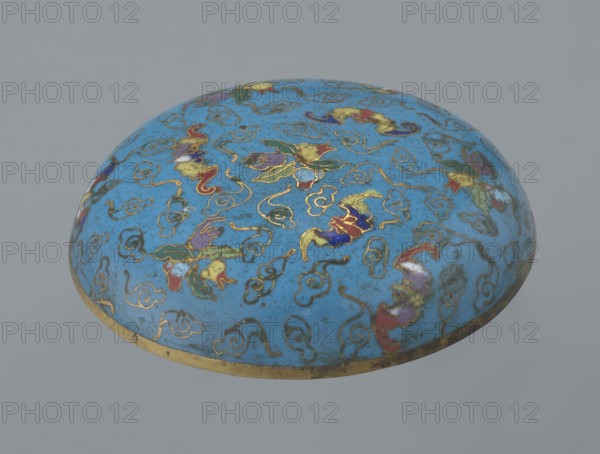 Round box with a lid, Chinese, Qing 1644–1911, cloisonné enamel, cylindrical flattened shape on a separate ring foot with a slightly convex lid, decorated overall with multi-colored lotus tendrils on a turquoise background. Lid shows three fruits peach, pomegranate, lemon with accompanying bats—an auspicious motif meaning many happiness, long life, and many sons. Asymmetrical composition set against a cloud background formed by bent wire; same motif appears in the box’s storage section, which is also decorated with colorful medallions single, pairs, or threes featuring various Chinese plant ornaments. Rounded edge of lid and storage section, lower edge of foot and its bottom are gilded. Bottom and entire interior covered with turquoise enamel. 19th century, China; container; height 4.6 cm, diameter 9.8 cm., A rounded dome-shaped object with a glossy blue surface decorated with a pattern of swirling line motifs and floral-like shapes outlined in metallic gold, with additional colors inc...