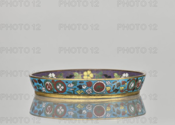 Plate., Gift; Paderewski, Ignacy Jan 1860–1941. Chinese; Qing dynasty 1644–1911. Cloisonné enamel decorative vessel huaniao style with images of birds swallows, plant motifs, flowers, floral ornaments, and plum trees. Plate with a brim, decorated on a turquoise background. Pair with a slightly different mirror decoration the companion plate differs. 19th century. Height 2 cm; diameter 14.2 cm., A shallow circular dish with a low rim sits on a reflective surface showing its mirrored underside; the dish has a bright turquoise exterior band decorated with a repeating pattern of circular medallions and small cloudlike motifs in colors including red, white, green, yellow, black, and gold, a thin gold band at the base, and a darker interior rim in purple with floral motifs in yellow, white, and green.