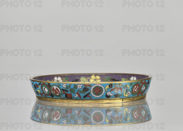 Plate., Chinese; Qing dynasty 1644–1911. Cloisonné enamel huaniao style. Decorative vessel with brim; turquoise background. Motifs: images of birds swallows, plum trees, flowers, and other plant/floral ornaments. Pair with a slightly differing mirror decoration. 19th century. Height 2 cm; diameter 14.2 cm., A low, shallow circular bowl displayed on a reflective surface, showing a colorful decorative band around its rim with a repeating pattern of circular medallions and abstract cloud- and floral-like motifs. The visible colors include turquoise, teal, blue, purple, red, green, yellow, white, black, and gold. The bowl has a gold-colored base rim and an inner surface with darker purple and floral shapes. The reflection beneath mirrors the patterned band and colors.