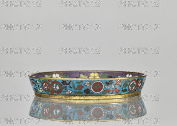 Plate. Cloisonné enamel decorative vessel, Plate with a brim. Turquoise-ground decoration with birds swallows, plant motifs, flowers, floral ornaments, plum trees; huaniao style; Qing dynasty 1644–1911, 19th century, China. Height 2 cm; diameter 14.2 cm. It is a pair with a slightly differing mirror-decoration counterpart., A shallow round dish with a low rim sits on a reflective surface, showing the dish's mirrored underside; the exterior rim features a bright turquoise background with decorative circular medallions and scrolling motifs in red, green, white, black, and gold, and a gold band runs along the base; the interior rim visible above shows a deep purple background with floral shapes in yellow, white, and green; the reflection repeats the turquoise, red, green, white, black, purple, yellow, and gold colors.