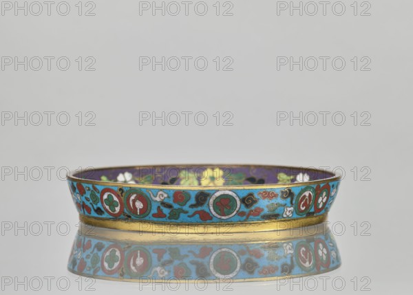 Plate. Cloisonné enamel huaniao, decorative vessel with a brim. Turquoise background with images of birds swallows, plant motifs, flowers, plum trees, and floral ornaments. Pair with a slightly different mirror decoration. 19th century, Qing dynasty 1644–1911, China. Height 2 cm; diameter 14.2 cm. Maker, inscription/gift: Paderewski, Ignacy Jan 1860–1941 noted., A shallow decorative bowl sits on a reflective surface, showing a mirrored reflection beneath it; the bowl has a bright turquoise band around its exterior adorned with circular and cloud-like motifs in red, white, green, black, and gold, and a gold rim and base; the interior visible at the top edge shows dark purple with floral patterns in yellow, green, white, and black; the background is a smooth pale gray.
