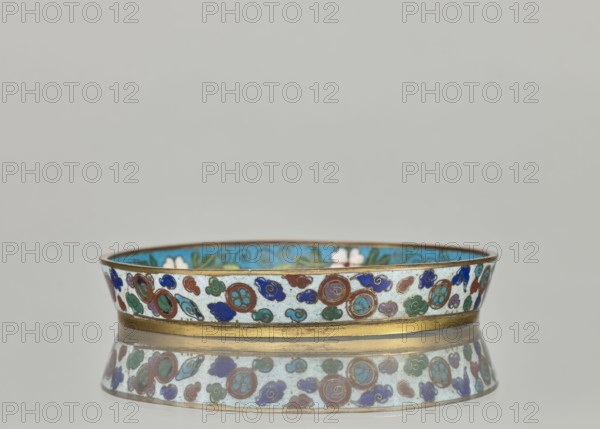 Plate with a brim; cloisonné enamel dish, 19th century China Qing dynasty. Circular plate with plant ornament and tiger symbol decoration. Height 2.4 cm; diameter 14.2 cm., A low, shallow circular dish with a metallic rim and base sits on a reflective surface, creating a mirrored image beneath it. The exterior band of the dish is decorated with a repeating pattern of rounded and cloud-like shapes in colors including blue, turquoise, green, red, and white, outlined with thin metallic lines. The interior visible along the top edge shows a bright turquoise background with floral and scroll motifs in green and white. The metallic rim and base appear brass or gold-toned, and the overall scene has a neutral light gray background.