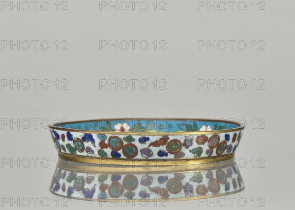 Plate, cloisonné enamel dish with brim, 19th century China Qing dynasty. Materials: cloisonné enamel. Decoration: plant ornaments and tiger motif symbol. Dimensions: height 2.4 cm, diameter 14.2 cm., A low, round decorative bowl with a thin gold rim and gold band near its base sits on a reflective surface, showing a mirrored image below. The exterior of the bowl features a repeating pattern of circular medallions and cloud-like motifs in colors including turquoise, blue, green, red, purple, and white against a pale background. The interior visible at the rim appears bright turquoise with floral shapes in white and green. The overall scene includes neutral gray background tones.