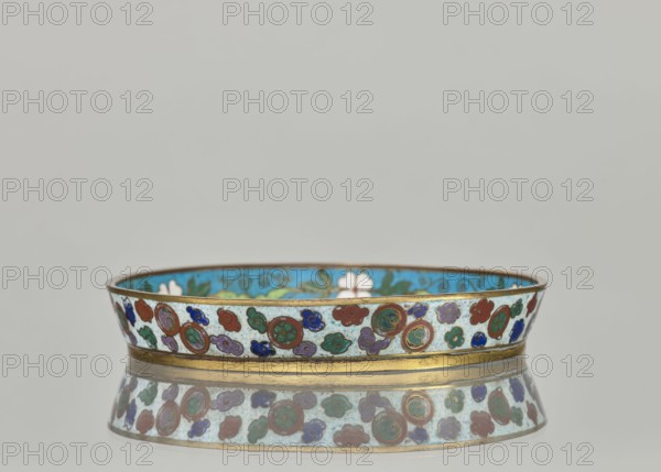 Plate, cloisonné enamel dish with a brim, 19th century China Qing dynasty. Height 2.4 cm; diameter 14.2 cm. Decorative vessel featuring plant ornaments and a tiger symbol. Maker, inscription/mark: author unspecified. : Paderewski, Ignacy Jan 1860–1941. Chinese art; Qing style., A shallow circular dish with a narrow flared rim sits on a reflective surface, showing a mirrored image below. The dish has a gold-colored outer band along the base and rim, and the exterior surface is decorated with a repeating pattern of round medallions and cloud-like shapes in red, green, blue, purple, and teal on a pale background. The interior visible near the rim is bright turquoise with stylized floral and vine motifs in green and white. The overall color palette includes gold, turquoise, red, green, blue, purple, teal, and pale off-white.