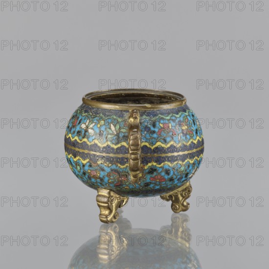 Ding incense burner, gift; Chinese, Qing dynasty 1644–1911; cloisonné enamel ceremonial tripod vessel, 18th century. Ding incense burner with two plastically shaped handles, no cover. Floral decoration in striped arrangements with floral motifs, butterflies, peonies, and fantasy beasts. Height 8.4 cm; width 12.4 cm; diameter 9.8 cm., A small, round metal vessel with a rounded body and an open top sits on a reflective surface; the body is decorated in a colorful cloisonné-style pattern featuring dominant shades of blue and turquoise with accents of yellow, red, green, white, and dark navy, and it has a horizontal wavy band encircling the middle in contrasting yellow and dark blue. The rim, handle, and three ornate feet are metallic gold in color; the handle is short and ribbed, attached vertically to the body, and the feet are curved with decorative scroll-like shapes. The interior appears dark and aged, and the vessel casts a soft reflection on the smooth, pale gray surface beneath it ...